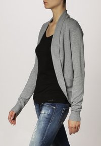 Cárdigan gris de punto con cuello de chal y detalles en canalé, combinado con una blusa negra y jeans azules desgastados, visto de lado.