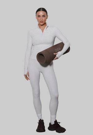 Kobieta w białym stroju sportowym trzyma zwiniętą brązową matę do jogi z logo, stojąc na jednolitym jasnoszarym tle.