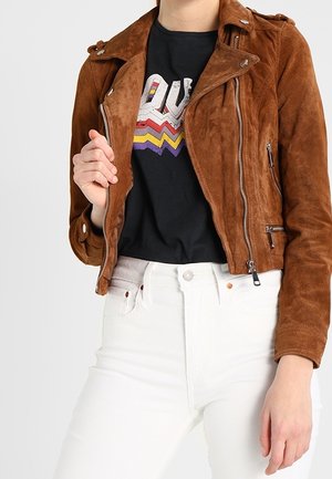 Personne portant une veste biker en daim marron sur un T-shirt graphique noir, associée à un jean blanc taille haute.