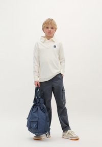 Cămașă polo bej cu mâneci lungi și guler, pantaloni cargo gri, adidași gri și albi, și un rucsac albastru cu șnur și buzunar frontal.