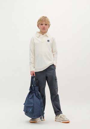 Beige lange mouwen polo shirt met een kraag, grijze cargo broek, grijs en witte sneakers, en een blauwe trekkoord tas met een voorvak.