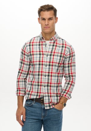 Man wearing a long-sleeve red, white, and blue plaid button-up shirt with rolled cuffs, blue jeans, and a wristwatch, standing with one hand in pocket.