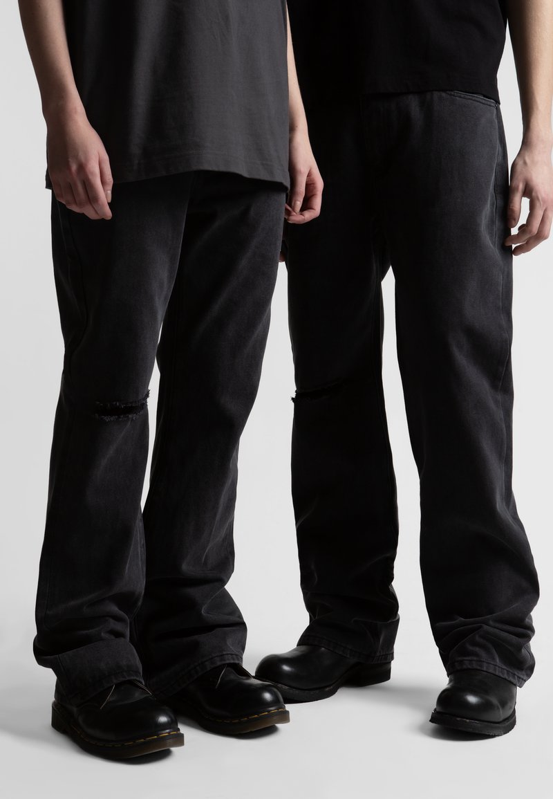 Δύο άτομα που φορούν μαύρα σκισμένα τζιν, μαύρα μπλουζάκια και μαύρα δερμάτινα παπούτσια, στέκονται κοντά σε απλό φωτεινό φόντο.