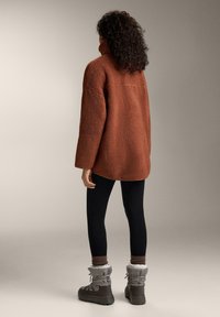 Rust-colored, textured fleece jacket with a high collar, paired with black leggings and gray winter boots featuring fur trim.