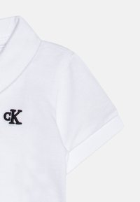 Vit polo t-shirt med en mjuk textur, korta ärmar och en svart broderad "CK" -logotyp på vänster bröst. Enkel och klassisk design.