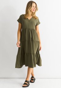 Olive green, knee-length dress with a V-neck, short sleeves, and tiered ruffles at hem. Paired with black sandals featuring gold accents.