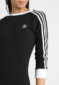 Svart långärmad tröja med vita tre-stripes detaljer på ärmarna och ribbade vita muddar, med en liten silverfärgad Adidas-logotyp på bröstet.