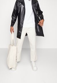 Sort, blank glasur regnfrakke, hvid t-shirt, cremede brede bukser og hvide platform sko. Holder en lys tote-bag med tekst.