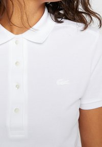 Polo blanc en fabric texturé, avec un col classique, une patte de boutonnage et un logo brodé discret sur la poitrine.