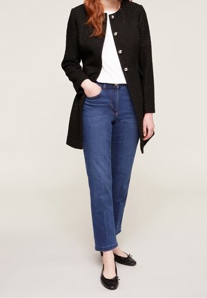 Schwarzer, strukturierter Mantel mit silbernen Knöpfen, weißem Oberteil, blauen geraden Jeans und schwarzen Ballerinas mit Schleifendetail.