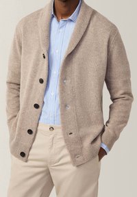 Beżowy kardigan z kołnierzem szalowym, prążkowanymi mankietami i sześcioma czarnymi guzikami; noszony na jasnoniebieskiej koszuli w paski i beżowych spodniach.
