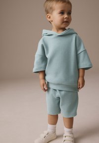 hele sinine tekstureeritud kapuutsiga dressipluus ja lühikeste pükstega komplekt, millel on kapuuts, lühikesed varrukad ja elastne vöökoht, kombineeritud valgete takjakinnitusega jalanõude ja sokkidega.