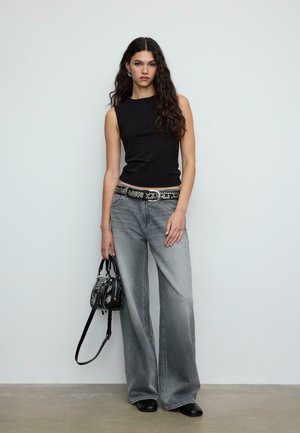 Top nero senza maniche con design aderente, abbinato a jeans grigi a gamba larga. Cintura nera con borchie e piccola borsa nera, su uno sfondo semplice.