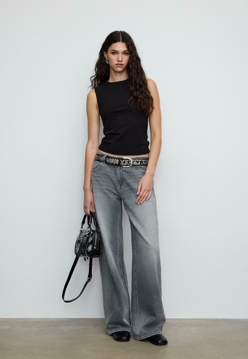 Czarny bezrękawnik o dopasowanym kroju, zestawiony z szerokimi szarawymi dżinsami. Czarny pasek z ćwiekami i mała czarna torebka, na jednolitym tle.