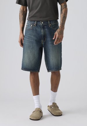 Person med tatoverede arme iført falmede blå denimshorts, hvide crew-sokker, lysebrune ruskinds slip-on sko og en mørkegrå t-shirt.