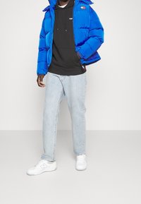 Giacca imbottita blu con cappuccio, felpa nera con coulisse sotto, jeans azzurri chiari e sneakers bianche. Presenta accenti del logo sulla giacca.