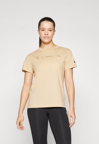 Beige tričko s krátkým rukávem vyrobené z bavlny s kulatým výstřihem, na přední straně je vyšívané logo "Champion" v kurzívě.