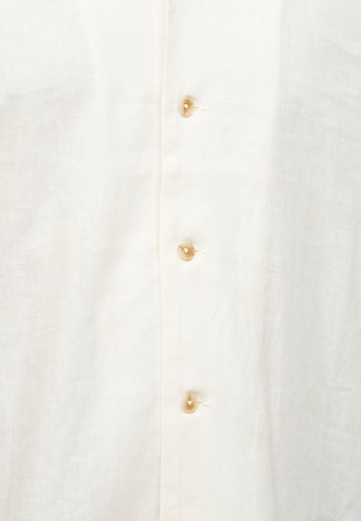 Κοντινό πλάνο σε κρεμ ύφασμα με τρία μπεζ κουμπιά ραμμένα κάθετα, που δείχνει την υφή και τις κουμπότρυπες σε ένα ελαφρύ ένδυμα.