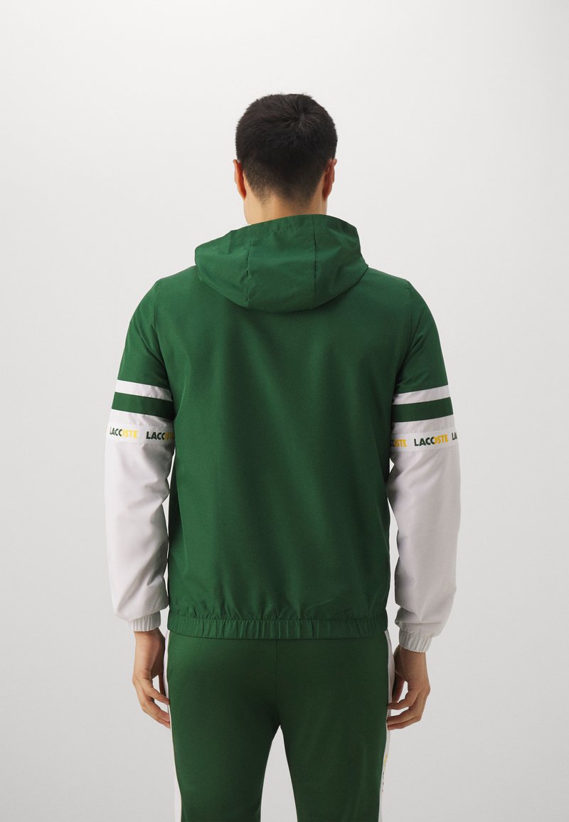 Hoodie mit grünem Körper und weißen Ärmeln, featuring horizontale Streifen und Logo-Akzente. Glatte Textur und elastischer Bund.