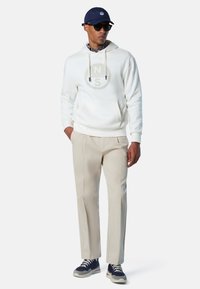 Felpa bianca con cappuccio e logo circolare, pantaloni beige su misura e sneaker blu navy. Il modello indossa occhiali da sole e un berretto blu navy.