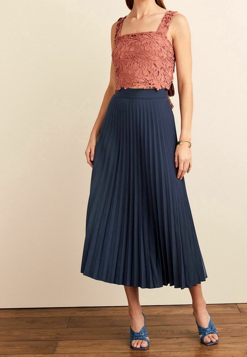 Femme portant un haut court en dentelle florale rose, une jupe midi plissée bleu marine, des talons ouverts bleus et un bracelet en or, debout sur un sol en bois.