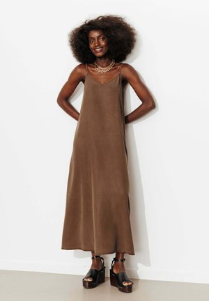 Femme avec une coiffure afro portant une robe longue marron sans manches et des sandales à plateforme noires, debout contre un mur blanc, souriante.