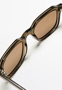 Kwadratowe okulary przeciwsłoneczne z półprzezroczystą ciemną oprawką, brązowymi szkłami i smukłymi metalowymi ramionami. Gładka powierzchnia i nowoczesne detale designu.