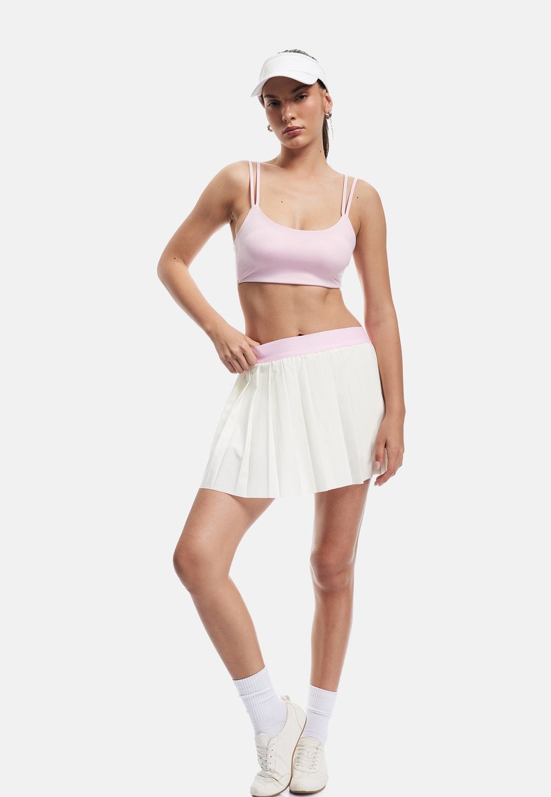 Lyserød crop top og hvid plisseret nederdel med en lyserød talje, parret med hvide sneakers og sokker, sat mod en ensfarvet baggrund.