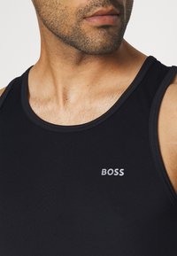 Černý top bez rukávů vyrobený z hladkého materiálu. Obsahuje kulatý výstřih a minimalistické logo "BOSS" v stříbrné barvě na přední straně. Jednoduchý design.