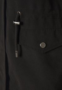 Černá látková kapsa s klopou zajištěnou kovovým patentem a připojenou černou šňůrkou s kovovým zarážkou na oděvu.