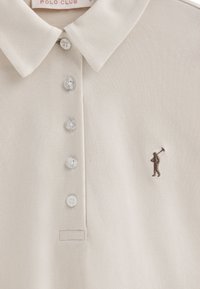 Kremowa koszulka polo o teksturowanej tkaninie, z kołnierzykiem, sześcioma perłowymi guzikami oraz brązowym haftowanym logo golfisty.