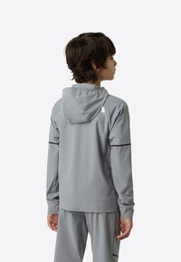 Grå hettejakke laget av lett stoff, med minimalistisk design, subtile svarte detaljer og en logo på baksiden.