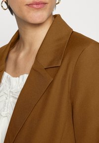 Brun blazer med en strukturerad krage och slät textur. Har en enkelknapprad och är kombinerad med en vit volangblus.