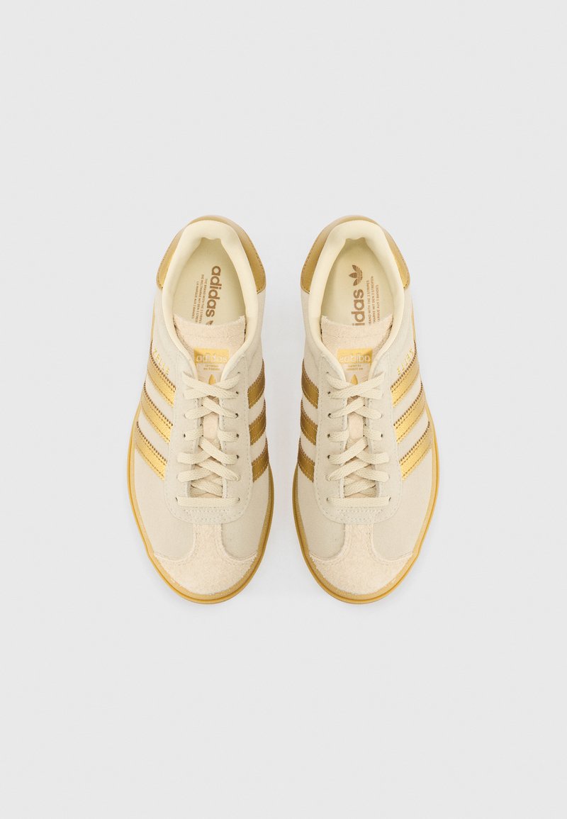 Beige Sportschuhe mit goldenen Streifen, aus Wildleder und Textilmaterialien, runde Spitze, flache Gummisohle und Schnürverschluss.