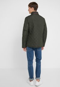 Quilted green jacket with a brown leather collar, featuring a tailored fit, side pockets, and a smooth texture. Paired with blue jeans and white sneakers.