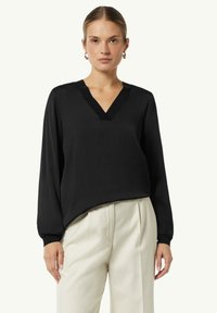 Schwarze Bluse mit langen Ärmeln und V-Ausschnitt, glatte Textur und leicht lockere Passform. Kombiniert mit hellen, taillierten Hosen.