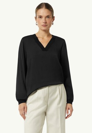 Schwarze Bluse mit langen Ärmeln und V-Ausschnitt, glatte Textur und leicht lockere Passform. Kombiniert mit hellen, taillierten Hosen.