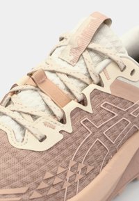 Zapatilla deportiva en rosa y crema con parte superior de malla texturizada, superposiciones de soporte, cordones tonales y un patrón geométrico distintivo en la suela.