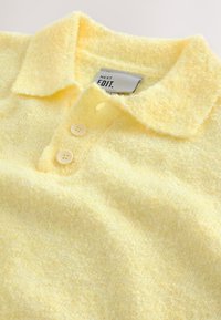 Polo jaune doux texturé avec deux boutons et une étiquette visible "NEXT EDIT." à l'intérieur du col.