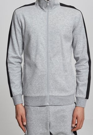 Homme portant une veste de survêtement gris clair à fermeture éclair avec des rayures noires le long des manches et un pantalon assorti gris clair.