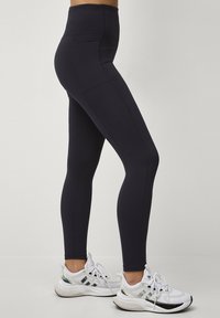 Højtaljede sorte leggings lavet af strækbart stof, med sidelommer og en glat tekstur, parret med hvide træningssko.