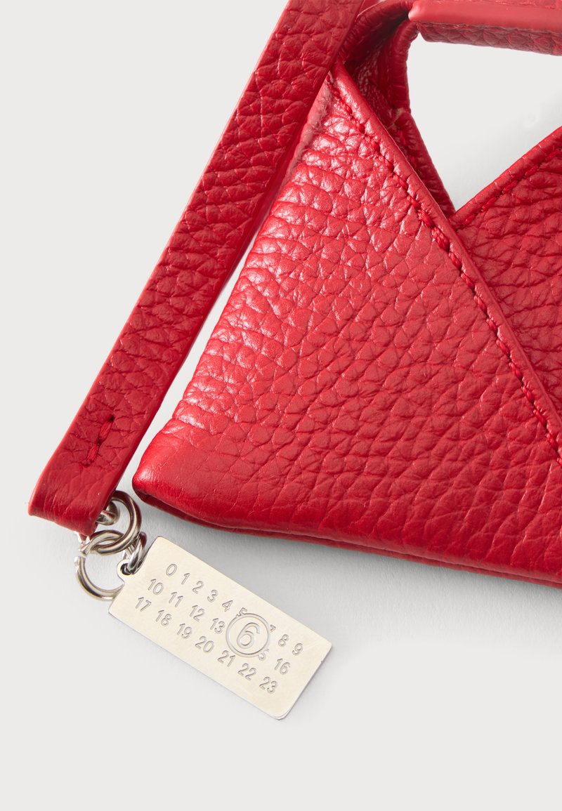 Gros plan sur le coin d'un sac en cuir rouge texturé avec une sangle et une étiquette métallique argentée attachée, gravée de chiffres et d'un six cerclé.