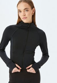 Chaqueta deportiva negra con cremallera y mangas largas, que presenta un diseño ajustado, tejido suave, cuello alto y agujeros para los pulgares en los puños.