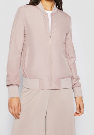 Femme portant une veste bomber rose clair avec fermeture éclair à l'avant et un pantalon ample assorti, debout devant un fond uni.