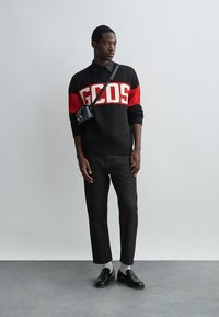 Juodas megztinis su raudonomis ir baltomis horizontaliomis juostelėmis, su "GCDS" logotipu, derinamas su tamsiomis džinsomis ir juodais odiniais batais.