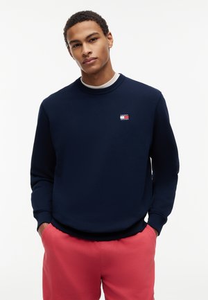 Νεαρός άνδρας που φοράει μπλε σκούρο φούτερ με μικρό λογότυπο και κόκκινο παντελόνι, στέκεται με τα χέρια στις τσέπες μπροστά σε λευκό φόντο.