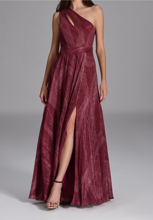 Femme portant une robe rouge longue jusqu'au sol, une épaule dénudée, avec une fente haute à la cuisse et des sandales à talons hauts à brides sur fond gris.