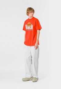 Camiseta roja de gran tamaño con un estampado gráfico de la cara de un oso y texto blanco, combinada con pantalones anchos blancos y zuecos beiges.