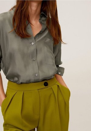 Mujer con una camisa suelta de botones de color verde oliva metida en unos pantalones plisados de tiro alto de color mostaza, con las manos en los bolsillos.