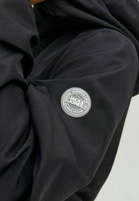 Černý látkový návlek s kulatou šedou nášivkou s nápisem „Jack & Jones“ a textem „Original Jeanswear Since 1980.“ Hladká textura.